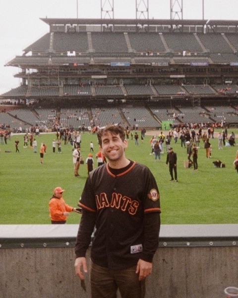 Me smiling in a black Giants jersey at the ballpark, with fans out on the field behind me