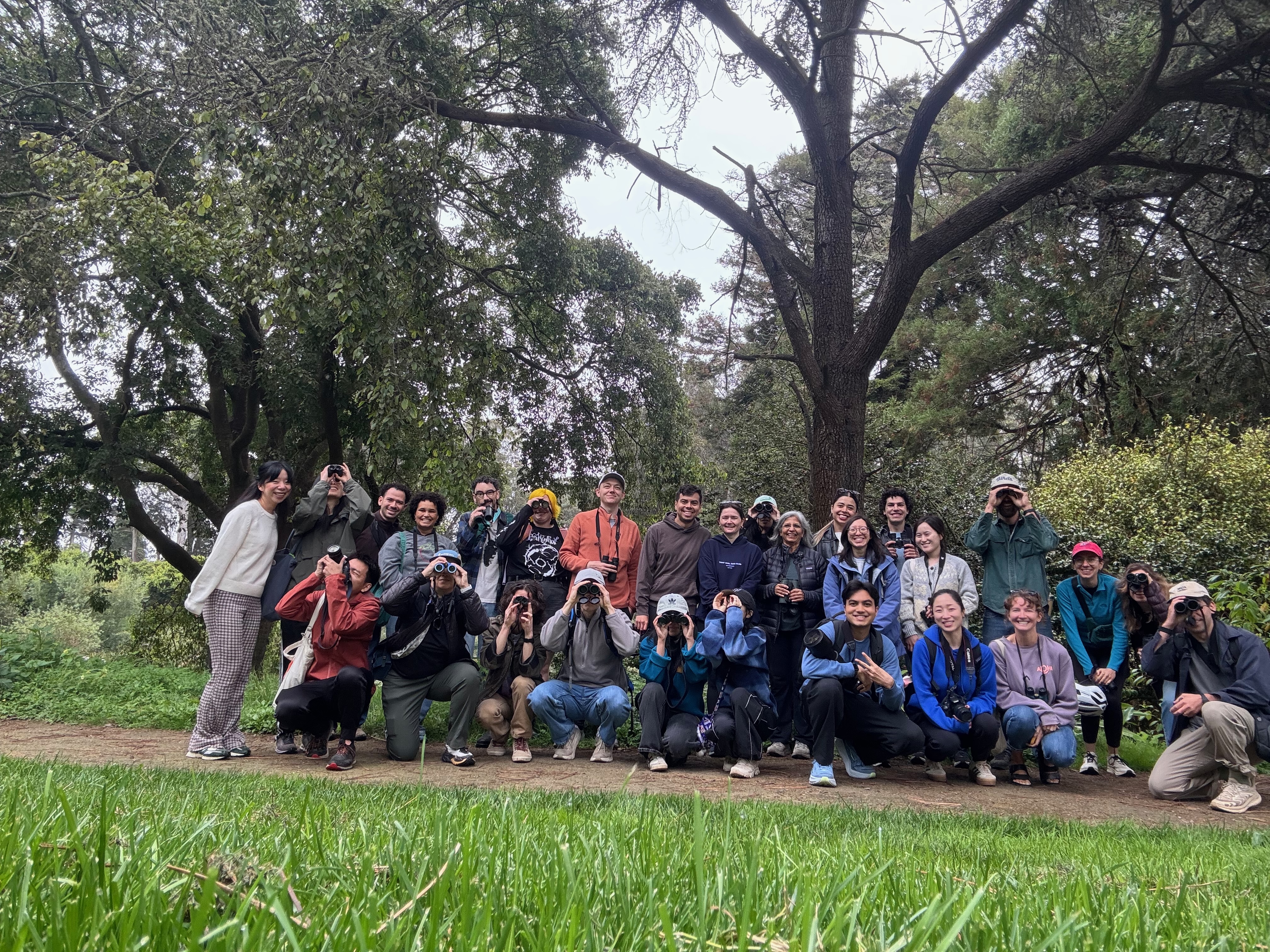 A large group of birders posing on a park trail, many holding binoculars up to their eyes